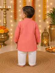A toddler standing with their back to the camera, wearing a coral pink kurta with thin vertical stripes and white pants, set against a festive decorated background with marigold garlands, string lights, and brass lamps.