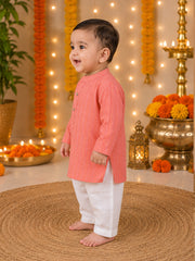 A toddler standing barefoot on a woven round mat, wearing a peach-pink kurta with white pants in a decorated festive indoor setting with marigold garlands, candles, and brass lanterns.