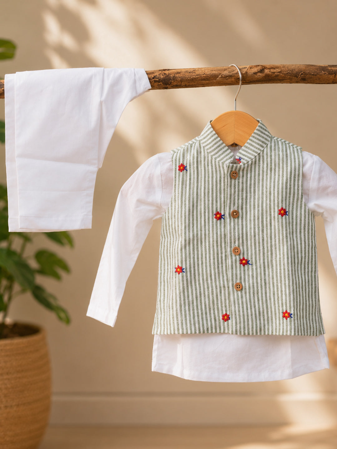 A children's outfit displayed on a wooden hanger, featuring a white long-sleeve shirt layered under a sleeveless vest with vertical green stripes and small red and blue embroidered details. A matching white fabric piece hangs from a wooden branch to the left, with a potted plant and warm beige wall in the background.