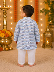 A toddler standing with their back to the camera, wearing a light blue and white patterned kurta with white trousers, set against a festive background with marigold garlands, a brass lamp, and a lantern.