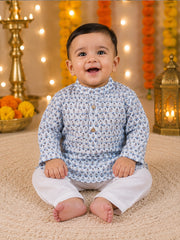A smiling baby sitting cross-legged on a beige carpet, wearing a light blue and white patterned kurta with white trousers, set against a festive background with marigold garlands and warm string lights.
