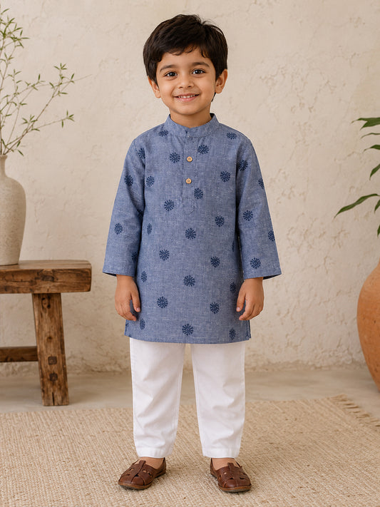 A young boy wearing a blue patterned kurta and white pants, standing in a styled indoor setting with a textured beige wall, a wooden stool, and a plant.