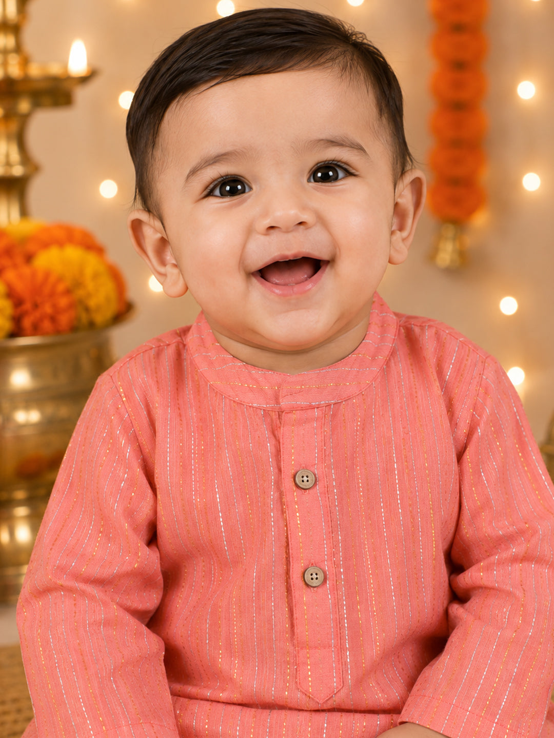 A smiling baby boy wearing a pink long-sleeve kurta with vertical metallic stripes, set against a warm festive background with marigold garlands and bokeh lights.